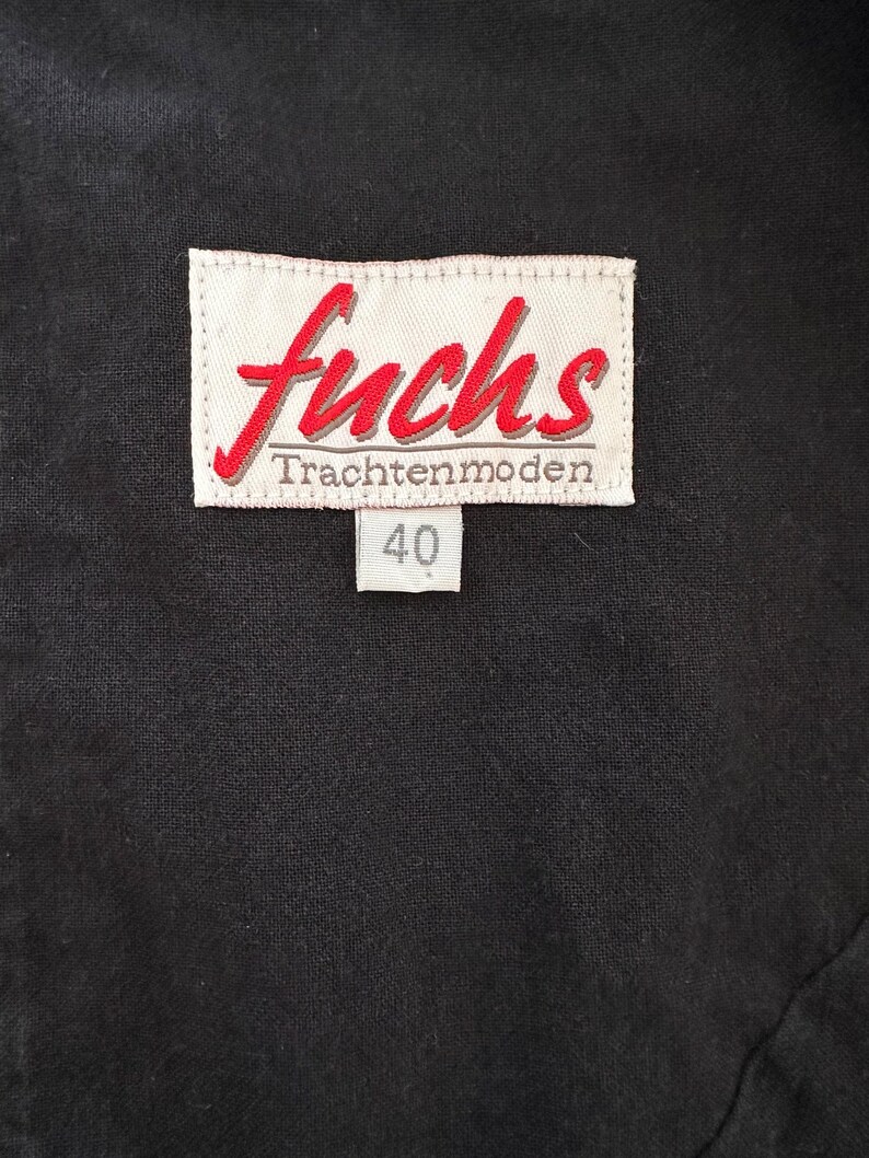 May include: Close-up of a black garment with a white label. The label features the red word "fuchs" in a cursive font, with "Trachtenmoden" below. A smaller tag displays the number "40".
