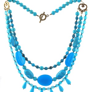 Op de afbeelding: Een drielaagse ketting in turquoise en blauw met een hartvormige sluiting en traanvormige kralen.