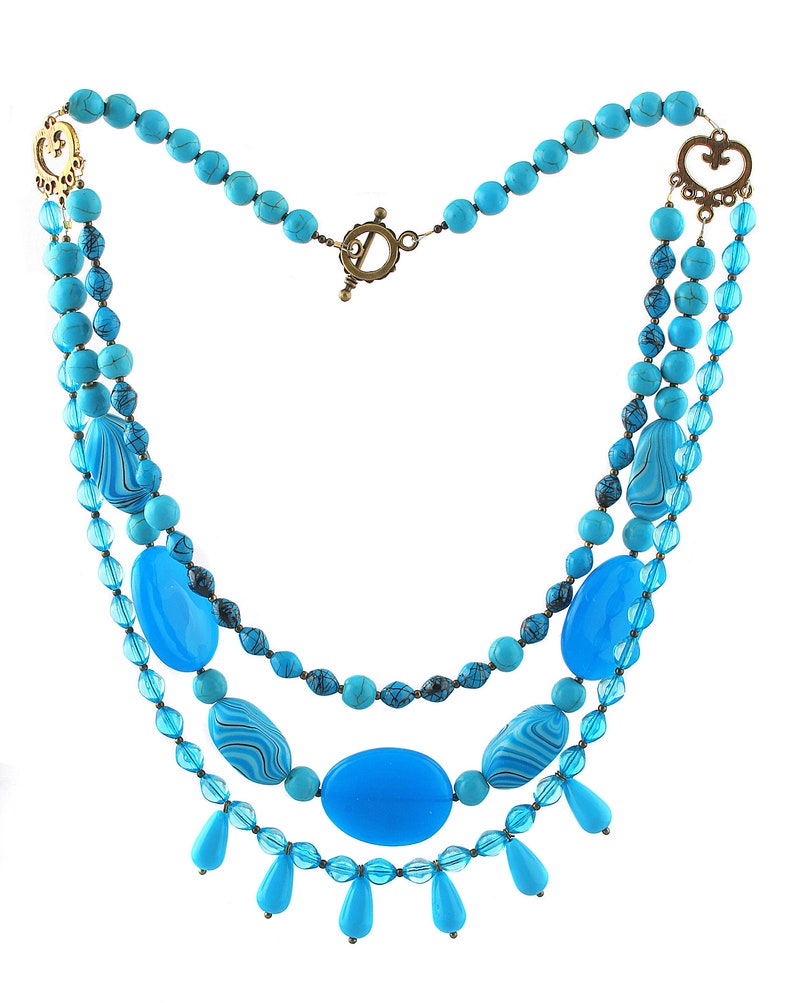 Op de afbeelding: Een drielaagse ketting in turquoise en blauw met een hartvormige sluiting en traanvormige kralen.