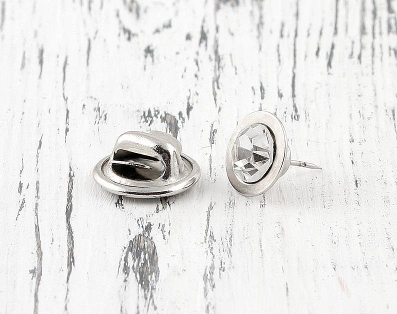 May include: Two silver-toned metal hat-shaped pin backs with a clear rhinestone in the center. The pin backs are used to attach a pin to a garment.