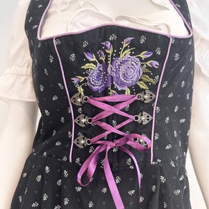 May include: Black dirndl with a floral pattern and purple embroidered roses. The bodice has a purple ribbon corset and silver clasps. The trim is purple, and the dress is worn over a white blouse.