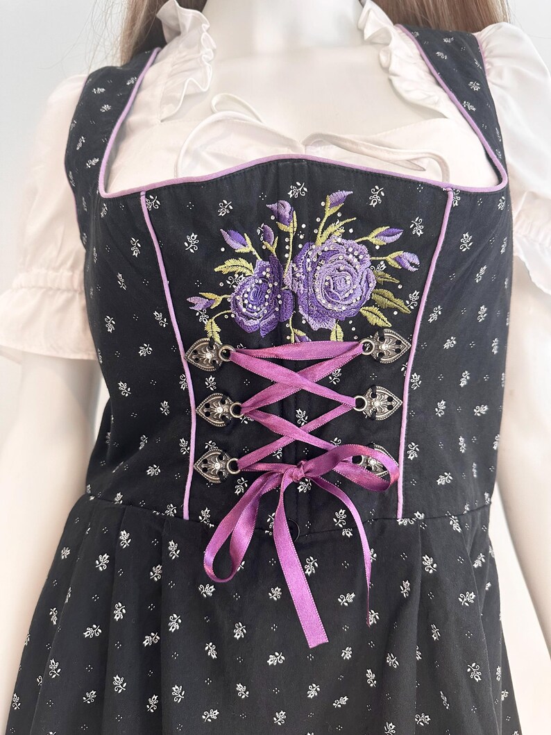May include: Black dirndl with a floral pattern and purple embroidered roses. The bodice has a purple ribbon corset and silver clasps. The trim is purple, and the dress is worn over a white blouse.