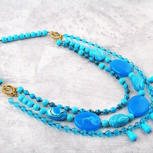 Op de afbeelding: Meerstrengs turquoise ketting met grote ronde en ovale kralen, sommige met een marmerpatroon. De ketting heeft een gouden sluiting en decoratieve accenten.