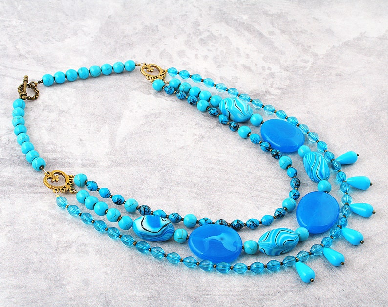 Op de afbeelding: Meerstrengs turquoise ketting met grote ronde en ovale kralen, sommige met een marmerpatroon. De ketting heeft een gouden sluiting en decoratieve accenten.