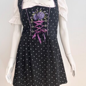 May include: Black dirndl dress with white floral pattern and purple accents. The dress features a square neckline, a bodice with purple embroidery, and a lace-up front. A white blouse with ruffled sleeves is visible underneath.