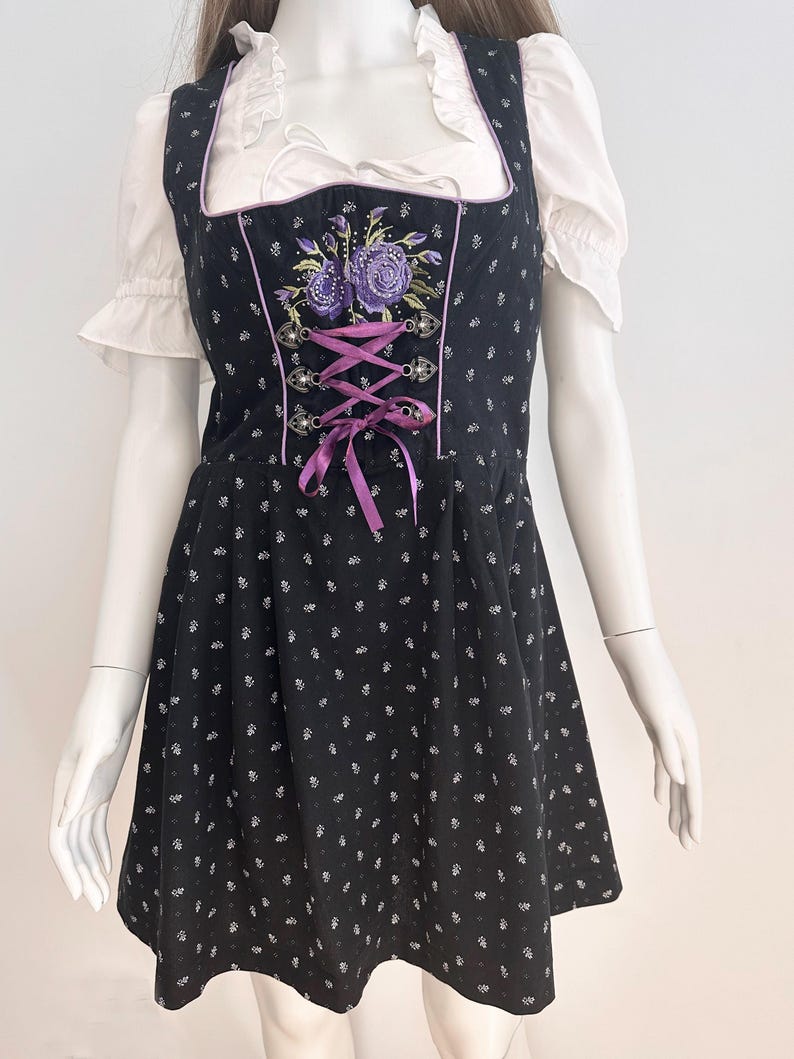 May include: Black dirndl dress with white floral pattern and purple accents. The dress features a square neckline, a bodice with purple embroidery, and a lace-up front. A white blouse with ruffled sleeves is visible underneath.
