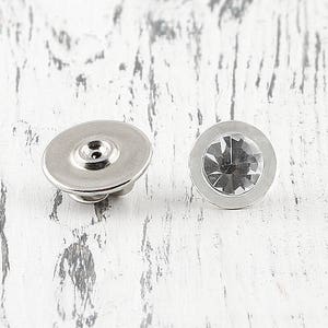 May include: Two silver metal button covers with a clear rhinestone in the center. The button covers are round and have a smooth surface.