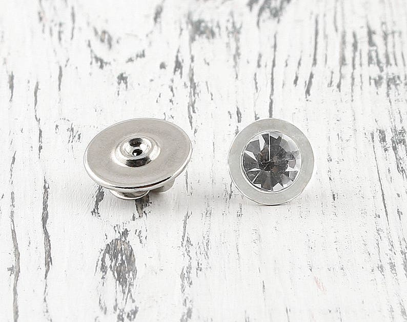 May include: Two silver metal button covers with a clear rhinestone in the center. The button covers are round and have a smooth surface.