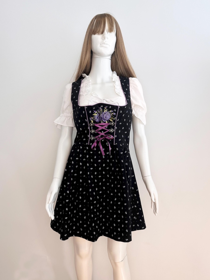 May include: Black dirndl dress with white floral pattern and purple accents. The dress features a square neckline, a bodice with purple lacing, and a white blouse with ruffled sleeves. The dress is displayed on a mannequin.