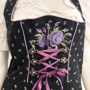 May include: Black dirndl bodice with a floral pattern and purple embroidered roses. The bodice has a pink trim, silver clasps, and a purple ribbon lace-up front. A white blouse with ruffled sleeves is visible underneath.
