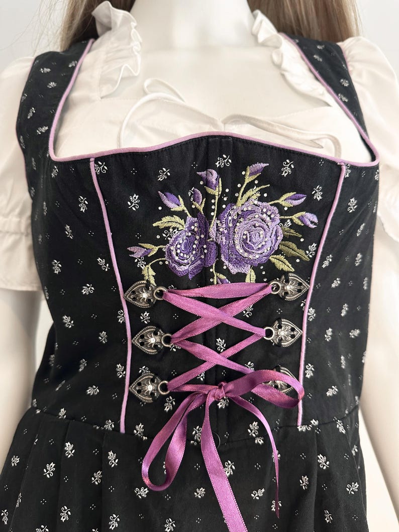 May include: Black dirndl bodice with a floral pattern and purple embroidered roses. The bodice has a pink trim, silver clasps, and a purple ribbon lace-up front. A white blouse with ruffled sleeves is visible underneath.