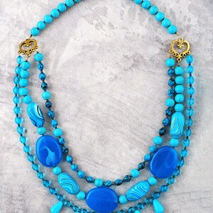 Op de afbeelding: Een gelaagde ketting in turquoise en blauw met een gouden sluiting en decoratieve accenten. De ketting heeft verschillende turquoise en blauwe kralen, waaronder ronde, ovale en druppelvormen.