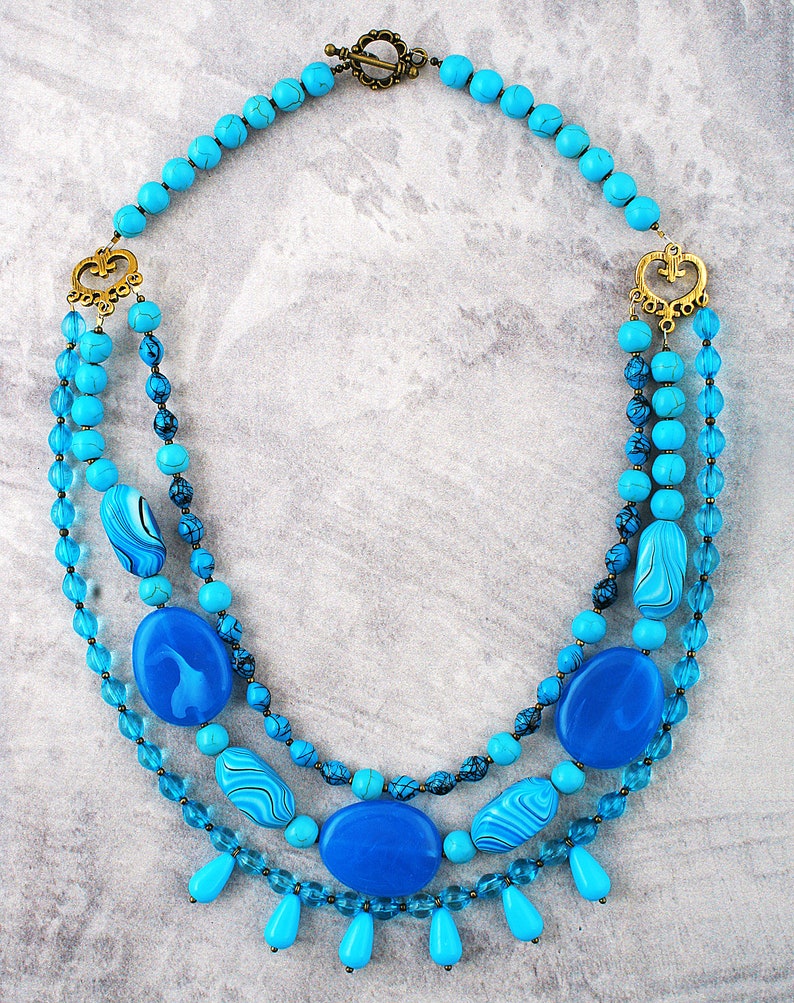 Op de afbeelding: Een gelaagde ketting in turquoise en blauw met een gouden sluiting en decoratieve accenten. De ketting heeft verschillende turquoise en blauwe kralen, waaronder ronde, ovale en druppelvormen.