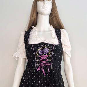 May include: Black dirndl dress with white floral pattern and purple embroidered flowers. The dress features a square neckline, white ruffled sleeves, and a purple ribbon corset. The dress is displayed on a mannequin.