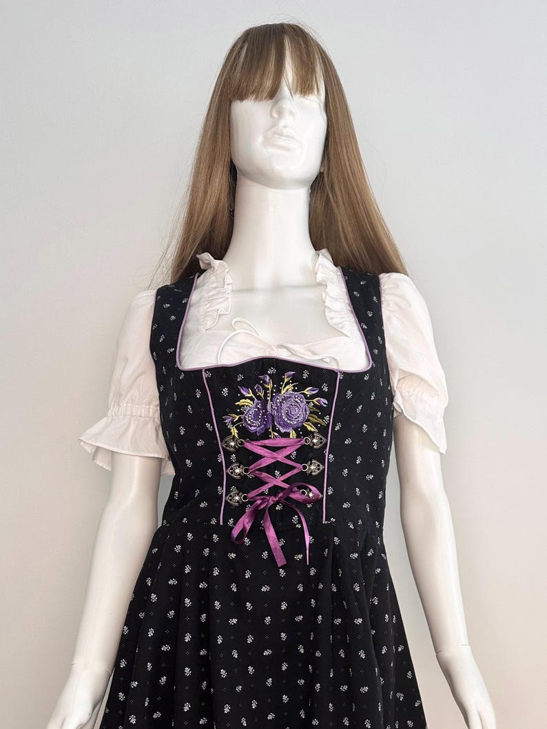 May include: Black dirndl dress with white floral pattern and purple embroidered flowers. The dress features a square neckline, white ruffled sleeves, and a purple ribbon corset. The dress is displayed on a mannequin.