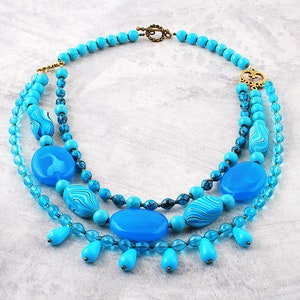 Op de afbeelding: Een gelaagde turquoise blauwe ketting met een gouden sluiting en grote turquoise blauwe kralen. De ketting heeft een traanvormig ontwerp met kleinere turquoise blauwe kralen.
