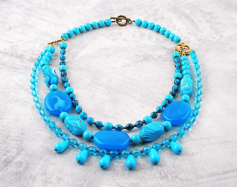 Op de afbeelding: Een gelaagde turquoise blauwe ketting met een gouden sluiting en grote turquoise blauwe kralen. De ketting heeft een traanvormig ontwerp met kleinere turquoise blauwe kralen.