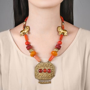 May include: A gold necklace with a large, ornate pendant featuring red stones. The necklace is made of orange, red, and yellow beads and has two gold lion figures on either side of the pendant.
