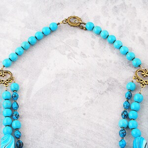 Op de afbeelding: Een turquoise en blauwe kralenketting met een gouden sluiting en decoratieve accenten. De ketting is gemaakt van meerdere strengen kralen en heeft een uniek ontwerp.
