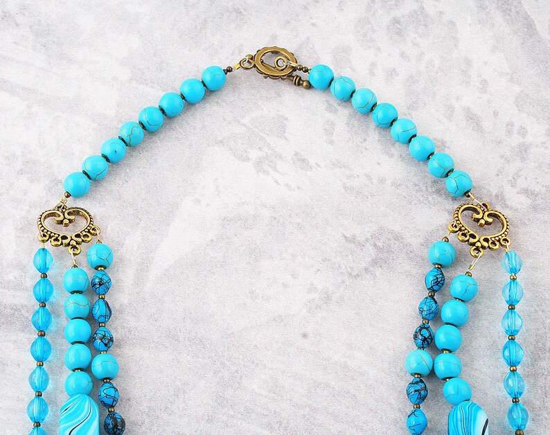 Op de afbeelding: Een turquoise en blauwe kralenketting met een gouden sluiting en decoratieve accenten. De ketting is gemaakt van meerdere strengen kralen en heeft een uniek ontwerp.