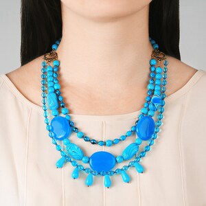 Op de afbeelding: Een meerrijige ketting met grote turquoise blauwe kralen en kleinere blauwe kralen. De ketting heeft een gouden sluiting en is gelaagd met kralen van verschillende groottes.