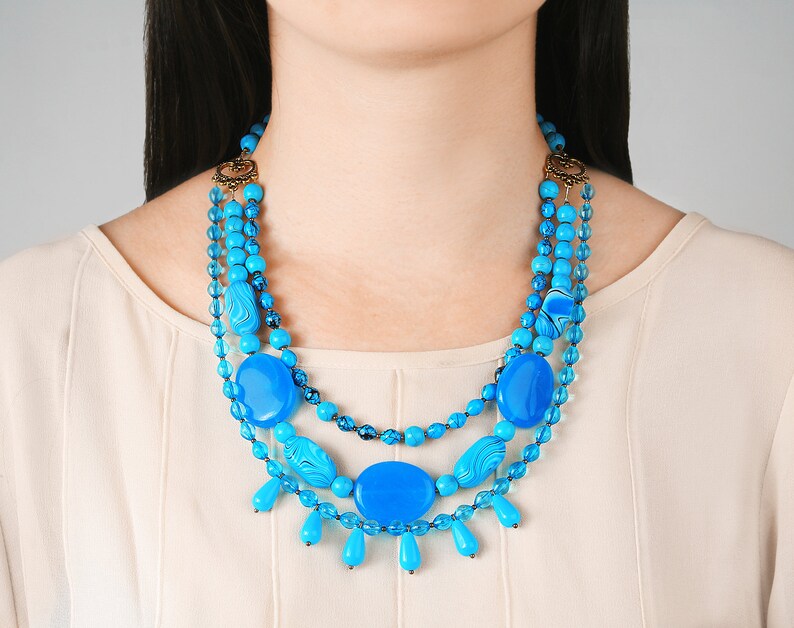 Op de afbeelding: Een meerrijige ketting met grote turquoise blauwe kralen en kleinere blauwe kralen. De ketting heeft een gouden sluiting en is gelaagd met kralen van verschillende groottes.