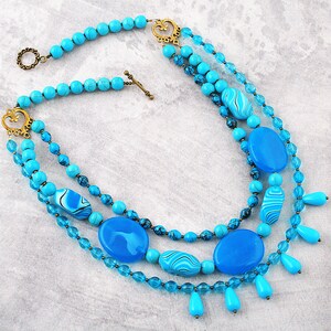 Op de afbeelding: Een meerrijige ketting met turquoise en blauwe kralen. De ketting heeft een verscheidenheid aan vormen en maten kralen, waaronder ronde, ovale en druppelvormen. De ketting is versierd met goudkleurige metalen sluitingen en connectoren.