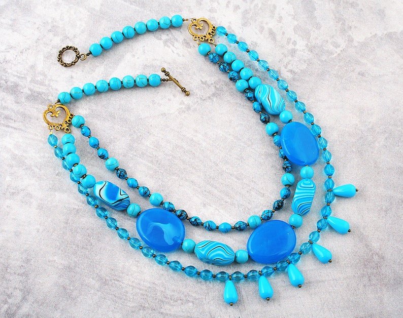 Op de afbeelding: Een meerrijige ketting met turquoise en blauwe kralen. De ketting heeft een verscheidenheid aan vormen en maten kralen, waaronder ronde, ovale en druppelvormen. De ketting is versierd met goudkleurige metalen sluitingen en connectoren.
