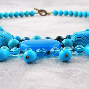 Op de afbeelding: Een turquoise en blauwe kralenketting met verschillende vormen en maten. De ketting heeft een mix van ronde, ovale en druppelvormige kralen.