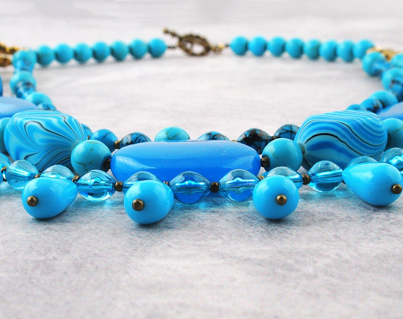 Op de afbeelding: Een turquoise en blauwe kralenketting met verschillende vormen en maten. De ketting heeft een mix van ronde, ovale en druppelvormige kralen.