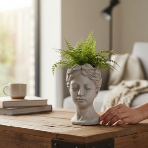 May include: A light grey stone-coloured planter shaped like a human head with detailed facial features and a wreath of leaves around the crown. A vibrant green fern plant is growing out of the top of the head, filling the opening of the planter.