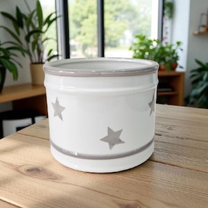 May include: White ceramic plant pot with a gray star design and gray trim. The cylindrical pot has a wide opening. The pot is suitable for indoor or outdoor use. The pot is on a wooden surface.