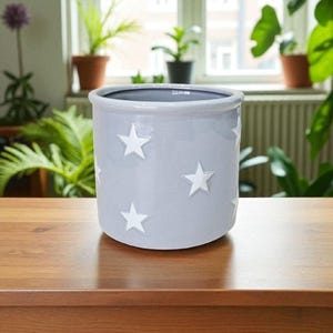 May include: A pale grey ceramic plant pot featuring white star designs. The pot is cylindrical and sits on a wooden surface. The background includes plants and a window. Suitable for indoor or outdoor use.