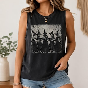 May include: A black sleeveless tank top featuring a graphic print of five figures wearing pointed hats and long dresses standing in a circle among trees. The text "Girls Will Be Girls." is printed in a rectangular box across the centre.