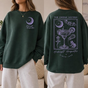 Könnte beinhalten: Ein dunkelgrünes Sweatshirt mit Rundhalsausschnitt, das vorne einen kleinen violetten Halbmond und den Schriftzug Midnight Margaritas zeigt. Auf der Rückseite befindet sich eine große rechteckige Grafik mit einem Margaritaglas, einer Fledermaus, einer Eidechse und einem Frosch.