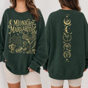 Könnte beinhalten: Ein dunkelgrünes Sweatshirt mit Rundhalsausschnitt und goldenem Grafikdruck. Die Vorderseite zeigt den Text Midnight Margaritas mit Abbildungen eines Frosches, eines Cocktailglases und einer Kaffeekanne. Die Rückseite zeigt eine vertikale Spalte.