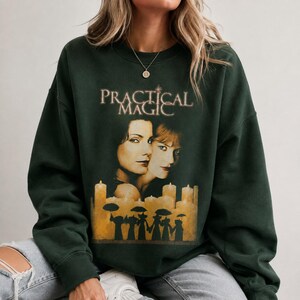 以下が含まれることがあります： ダークフォレストグリーンのクルーネックトレーナー。映画タイトル「Practical Magic」がセリフ体でプリントされています。テキストの下には2人の女性の顔と、傘を持った人物のシルエットが描かれた、灯ったキャンドルの列がデザインされています。