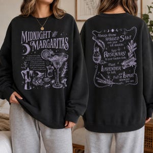 May include: A black crewneck sweatshirt featuring a light purple graphic print. The front displays the text Midnight Margaritas with a cocktail glass, a frog, and a coffee pot. The back features text about salt, rosemary, lavender, and love.