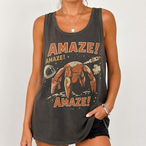 May include: A charcoal gray sleeveless tank top featuring a graphic print of a robotic creature, an astronaut helmet, and a rocket ship. The text AMAZE! is printed in orange above and below the central illustration, set against a starry background.