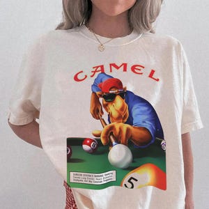 Op de afbeelding: Cr&egrave;mekleurig T-shirt met een afbeelding van een kameel die pool speelt, een zonnebril en een rode pet draagt. Het woord "CAMEL" is in het rood boven de afbeelding gedrukt. Het shirt bevat ook een waarschuwing van de Surgeon General.