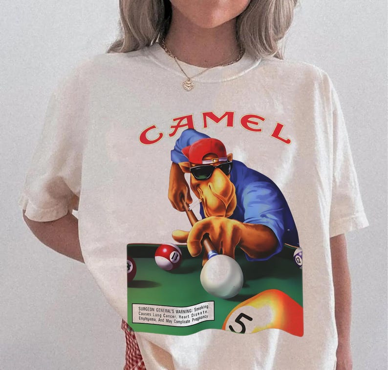 Op de afbeelding: Cr&egrave;mekleurig T-shirt met een afbeelding van een kameel die pool speelt, een zonnebril en een rode pet draagt. Het woord "CAMEL" is in het rood boven de afbeelding gedrukt. Het shirt bevat ook een waarschuwing van de Surgeon General.