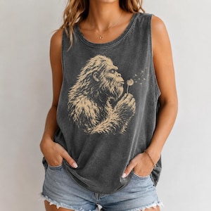 Puede incluir: Una camiseta sin mangas de color gris carbón con un estampado gráfico en beige de una criatura peluda soplando un diente de león. La prenda tiene una textura de tela desgastada, cuello redondo y tirantes anchos para un ajuste holgado.