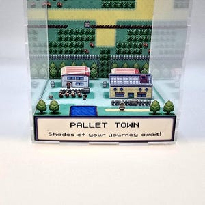 May include: A 3D diorama of Pallet Town from a video game, featuring two buildings, trees, and a pool. The text "PALLET TOWN" and "Shades of your journey await!" are displayed on a sign at the bottom.