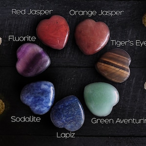 May include: Heart-shaped gemstones in red jasper, orange jasper, fluorite, tiger's eye, sodalite, lapis, and green aventurine are arranged in a circle. The names of the stones are written in white text. The background is a dark wood surface with white flowers.