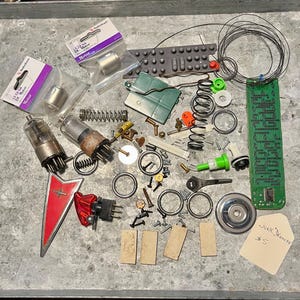 May include: A collection of electronic components and hardware, including vacuum tubes, springs, wires, and small motors. A red and white emblem and a green circuit board are also present. A tag reads "Junk Drawer #5".