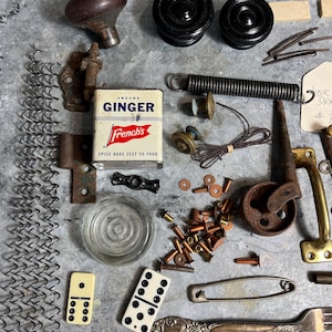 May include: An assortment of vintage hardware and small objects, including door knobs, wheels, springs, a spice tin, dominoes, and a fork, arranged on a metal surface. The French's Ginger tin is visible. The items are in various colors, including brown, black, and brass.