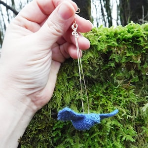 Può includere: Un ciondolo a forma di uccello blu all'uncinetto appeso a un anello di filo d'argento. Il ciondolo è sospeso davanti a uno sfondo verde muschio.