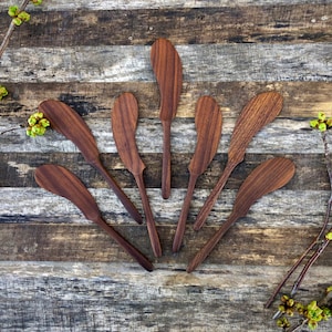 Puede incluir: Siete cuchillos de mantequilla de madera dispuestos en forma de abanico sobre un fondo de madera rústico. Los cuchillos son todos de madera marrón oscura y tienen un mango liso y redondeado.