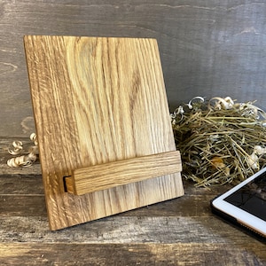 Peut inclure: Un support de téléphone en bois avec une finition en bois naturel. Le support a une barre horizontale pour maintenir un téléphone ou une tablette en place. Le support est posé sur une surface en bois.