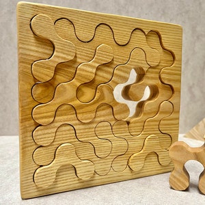 Könnte beinhalten: Ein Holzpuzzle mit einem einzigartigen ineinandergreifenden Design. Das Puzzle ist aus hellbraunem Holz gefertigt und hat eine quadratische Form. Dem Puzzle fehlt ein Teil, das separat gezeigt wird. Das fehlende Teil ist ebenfalls aus hellbraunem Holz gefertigt und hat eine einzigartige Form.